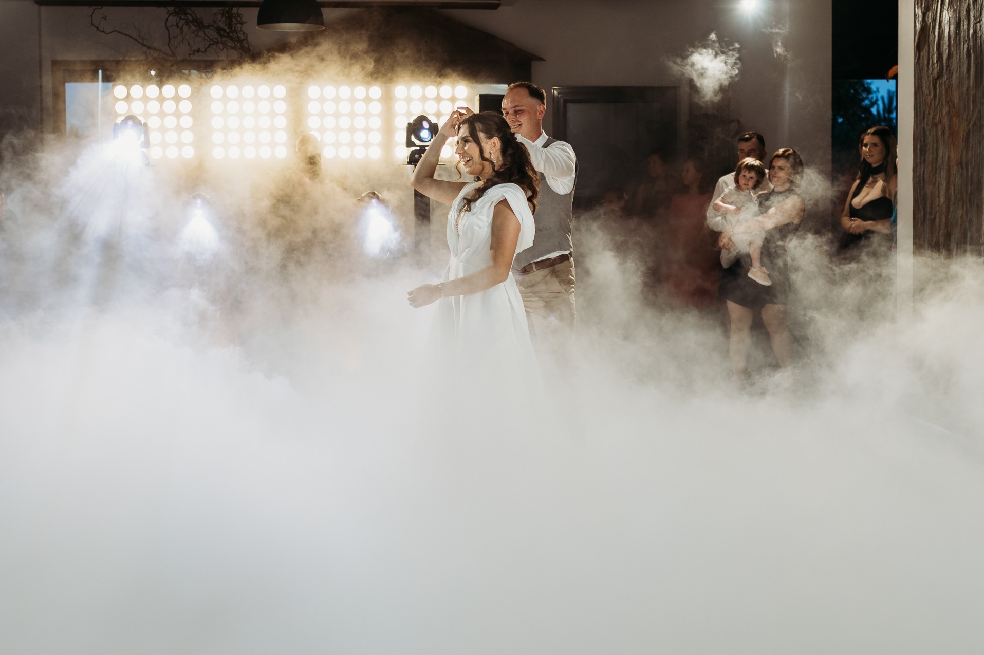 Para młoda tańczy w dymie na parkiecie, w tle oświetlenie sceniczne i goście. Ujęcie z boku, z widocznym efektem dymu i światła tworzącym unikalny klimat.