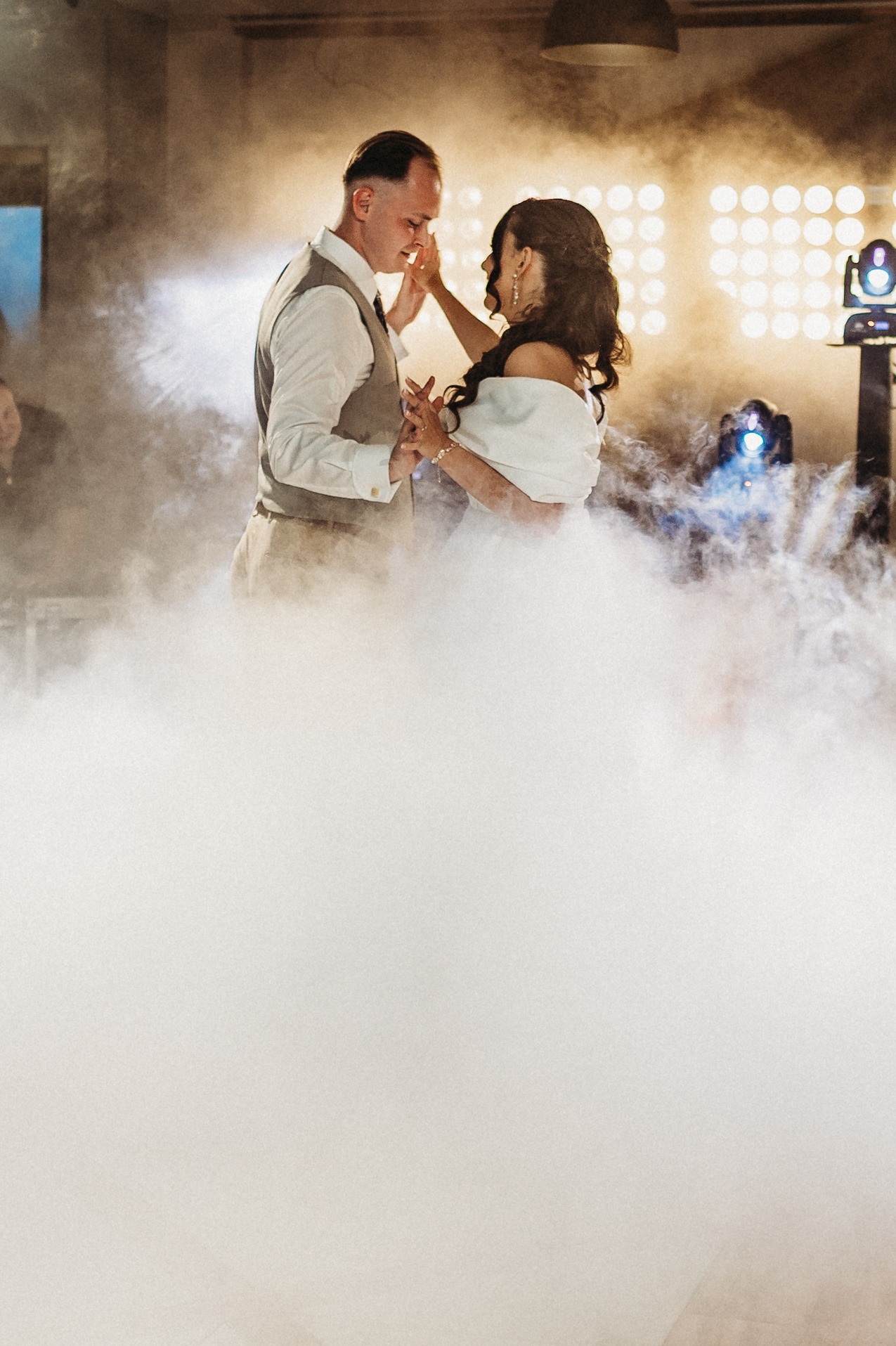 Para młoda tańczy w dymie na tle świateł scenicznych. Pan młody w kamizelce, panna młoda w białej sukni. Romantyczna atmosfera, oświetlenie eventowe.
