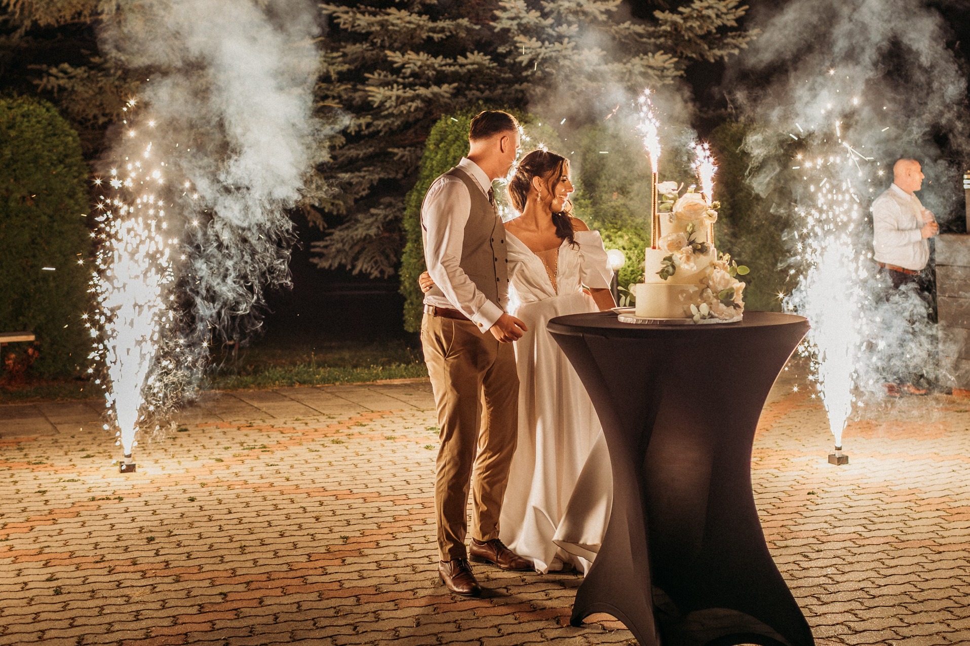 Uśmiechnięta para młoda stoi obok tortu weselnego, otoczona fontannami iskier na tle zieleni. Mężczyzna w brązowych spodniach i kamizelce obejmuje kobietę w białej sukni.