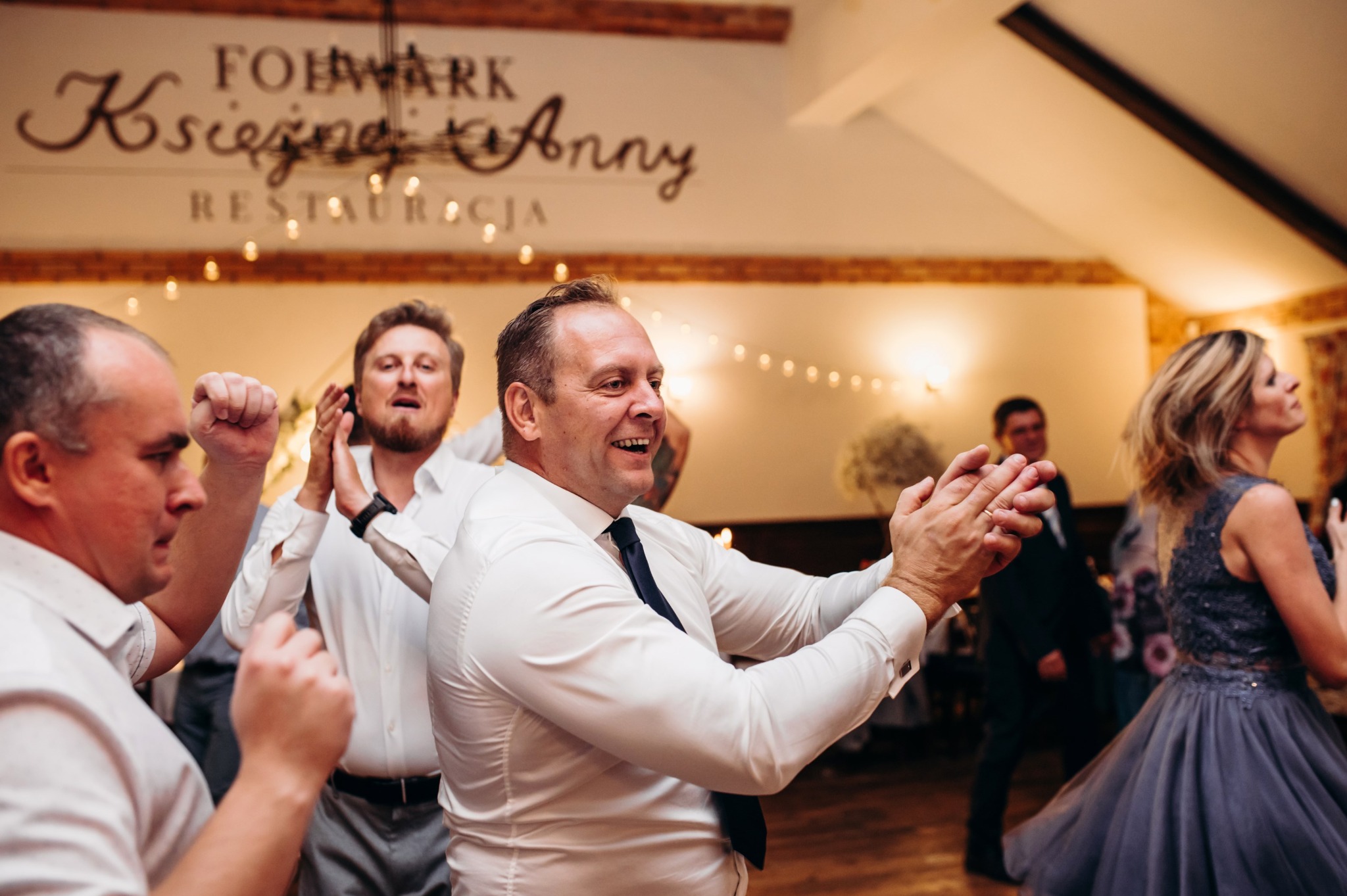Rozbawieni goście weselni w trakcie zabawy tanecznej w sali bankietowej z dekoracjami świetlnymi i napisem 'Folwark Księżnej Anny Restauracja'.