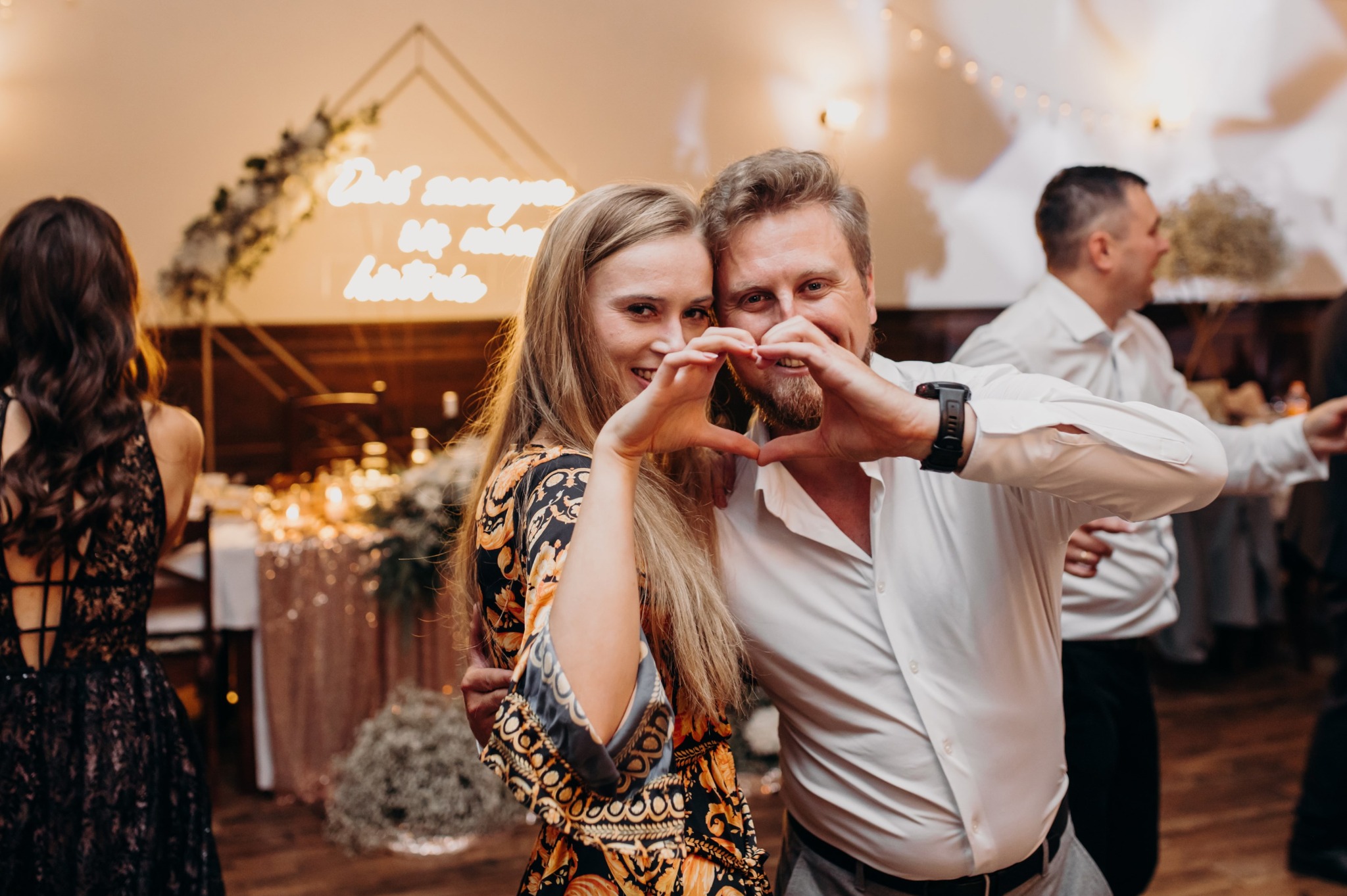 Uśmiechnięta para tworzy serce dłońmi na tle dekoracji ślubnej z neonowym napisem i girlandami świetlnymi. Parkiet i goście w tle.