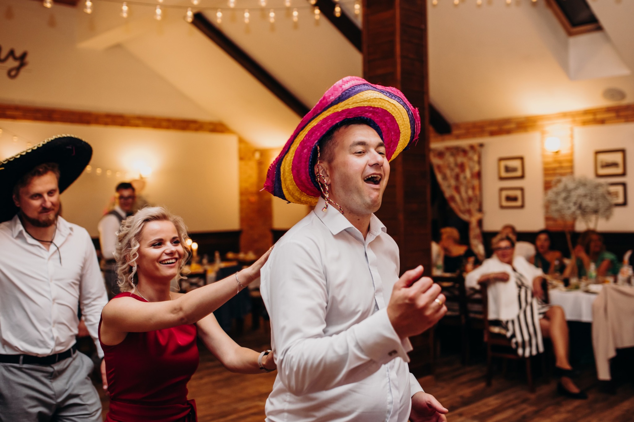 Rozbawieni goście weselni w kolorowych sombrerach tańczą na parkiecie. W tle widoczne stoły z gośćmi i dekoracje sali.