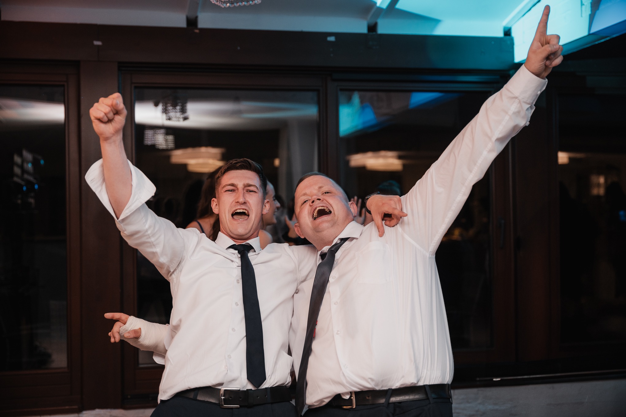 Dwóch mężczyzn w białych koszulach i krawatach, z uniesionymi rękami, świętuje na tle okien i oświetlenia imprezowego. Jeden z mężczyzn ma bandaż na dłoni.