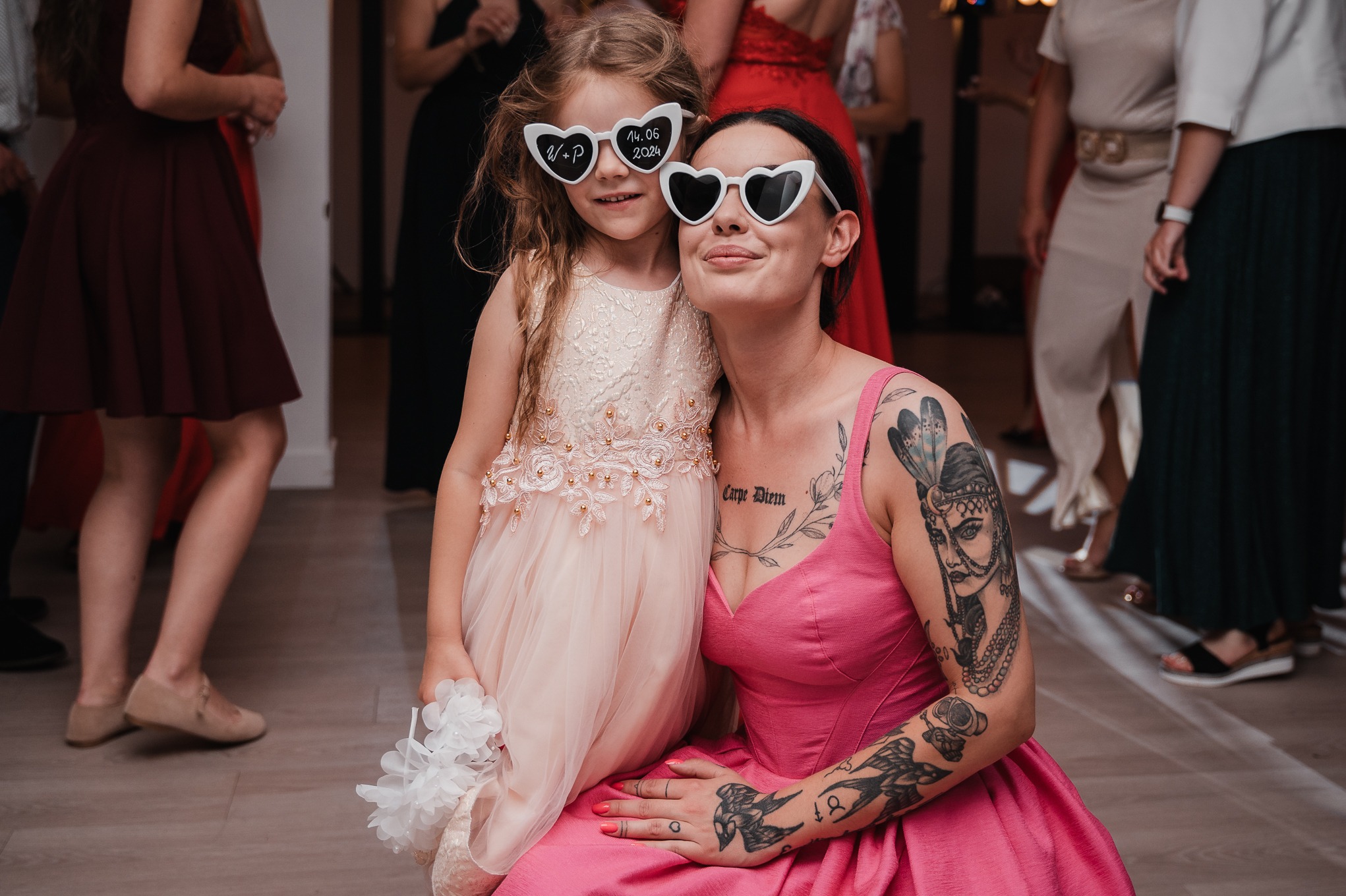 Uśmiechnięta dziewczynka w okularach w kształcie serc z datą ślubu, przytulona do kobiety w różowej sukience z tatuażami, na tle gości weselnych.