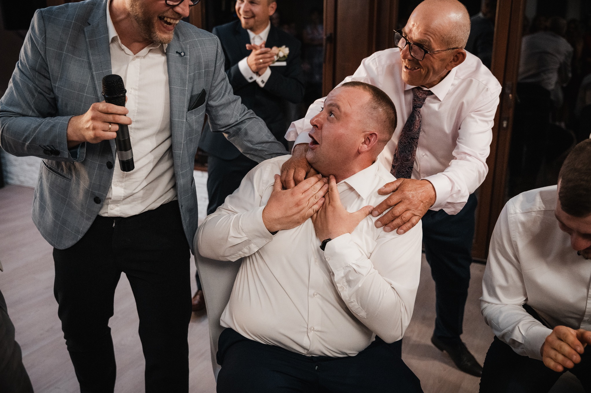 Rozbawiony mężczyzna z mikrofonem w dłoni, w otoczeniu gości weselnych, w trakcie zabaw oczepinowych. Uśmiechnięte twarze, jasne oświetlenie.