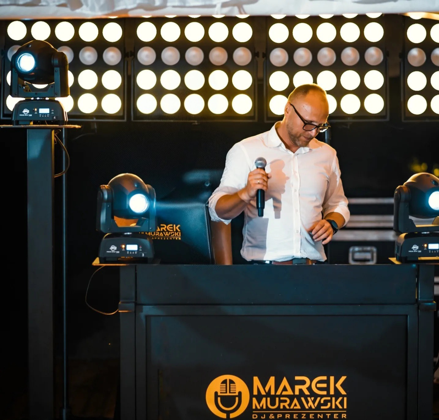 DJ Marek Murawski za konsoletą z mikrofonem, otoczony oświetleniem scenicznym. Widoczne logo firmy na froncie konsolety. Ujęcie z dołu.