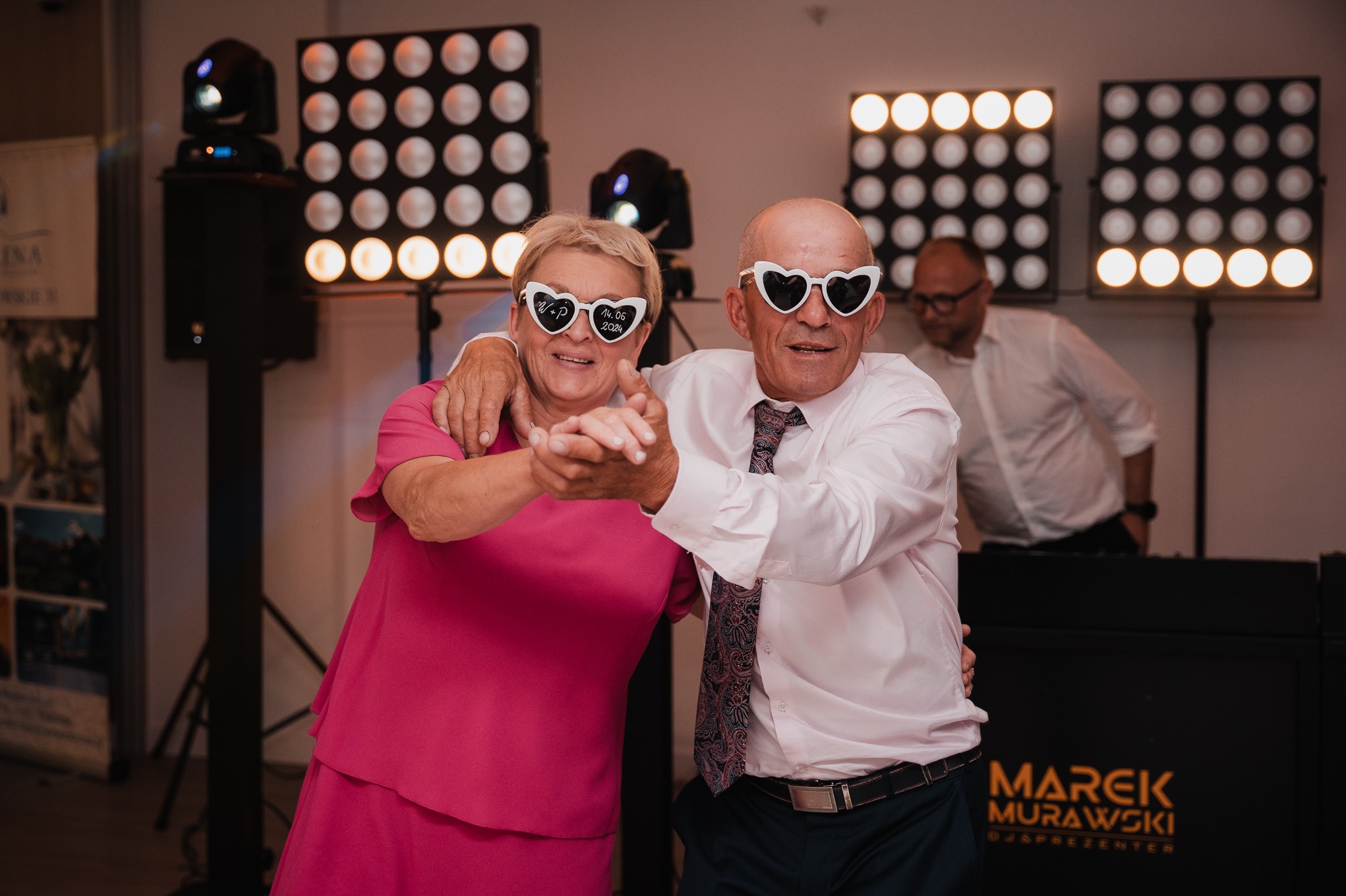 Para w okularach w kształcie serc tańczy na tle oświetlenia eventowego. Widoczny DJ Marek Murawski. Ujęcie z imprezy, prawdopodobnie wesela.