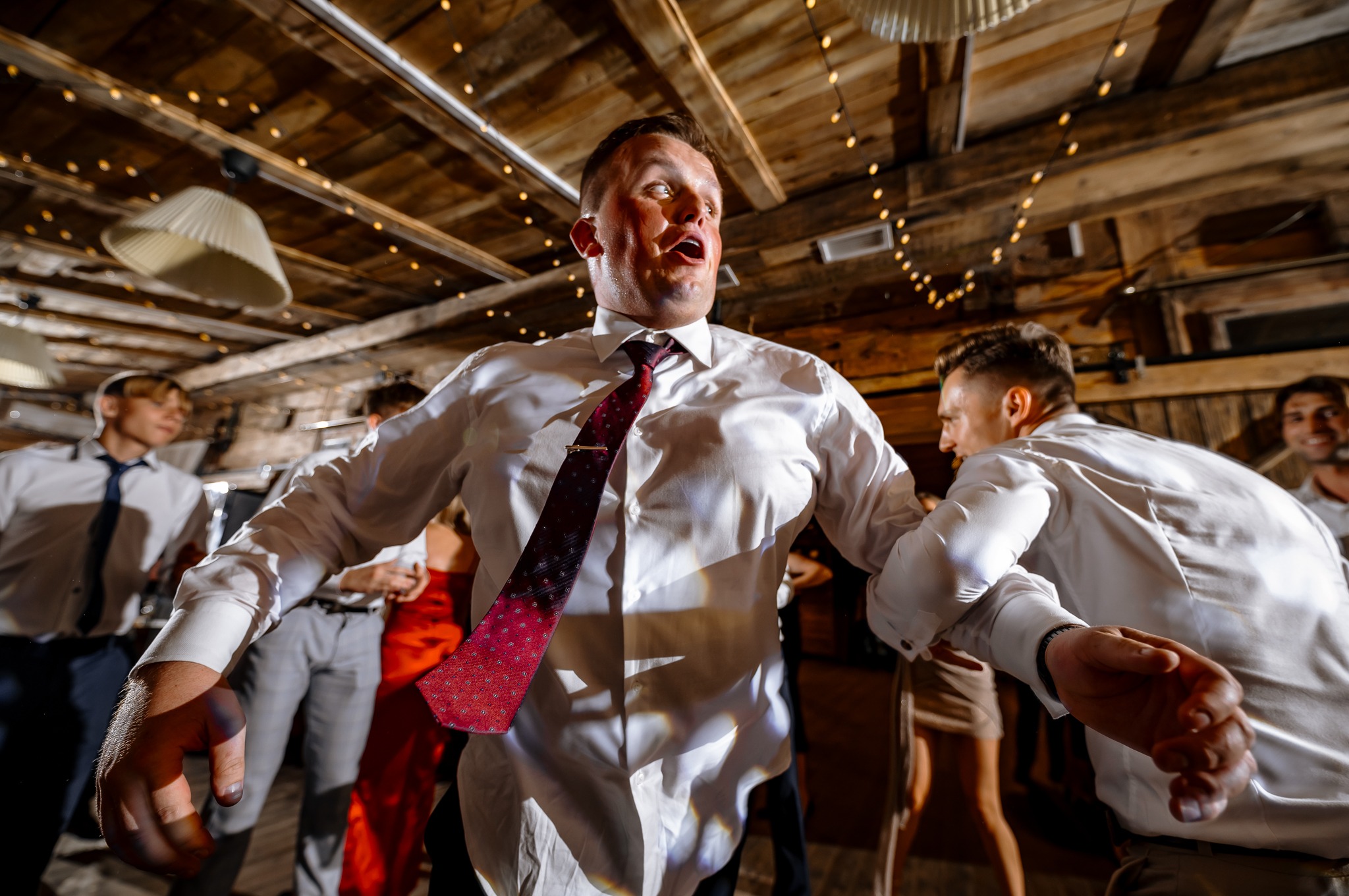 Rozbawiony mężczyzna w białej koszuli i bordowym krawacie tańczy na parkiecie podczas imprezy. W tle drewniane ściany i dekoracyjne oświetlenie.