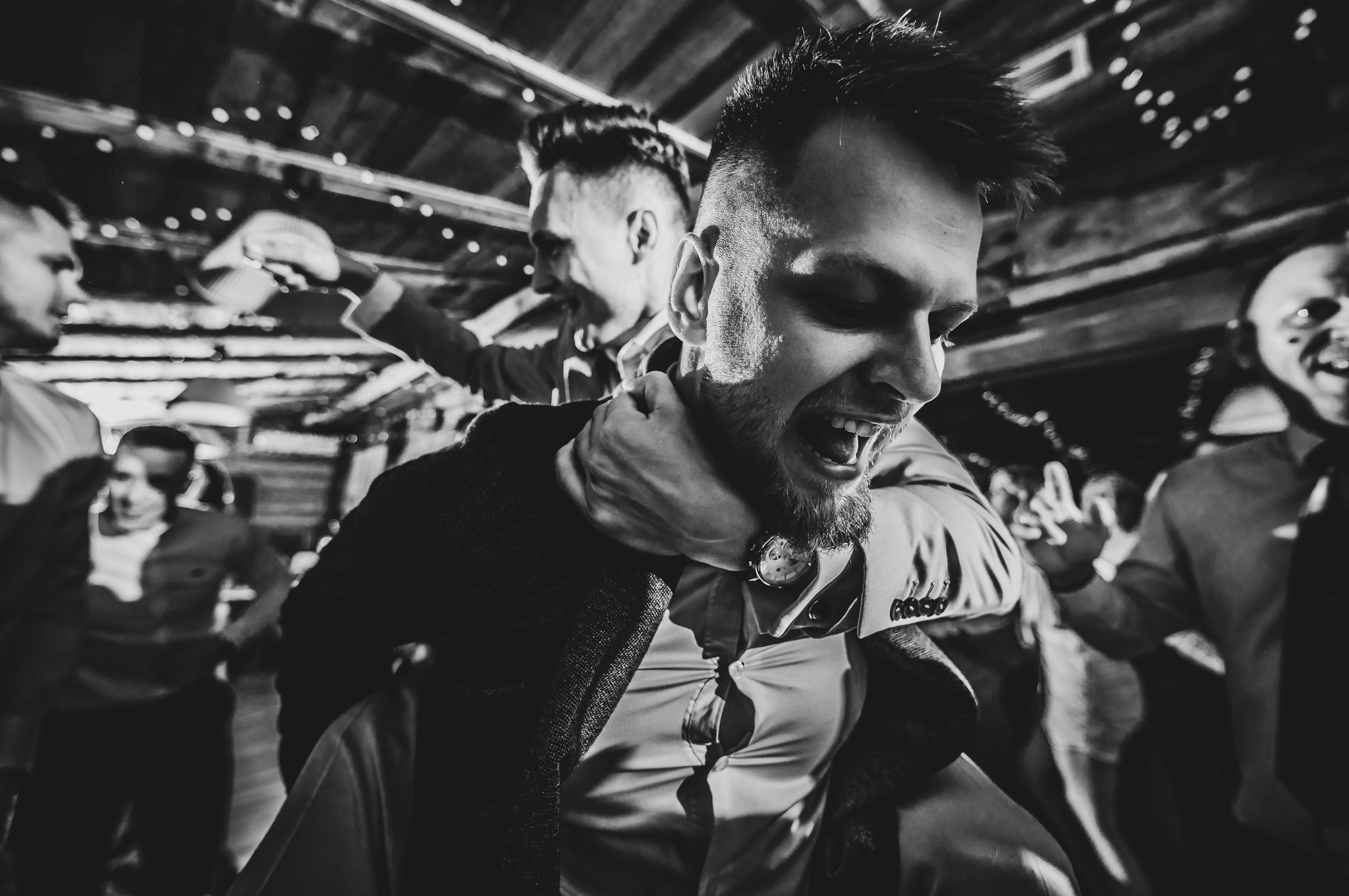 Czarno-białe zdjęcie rozbawionego mężczyzny niesionego na barana podczas imprezy. W tle widoczni inni uczestnicy zabawy i dekoracyjne oświetlenie.