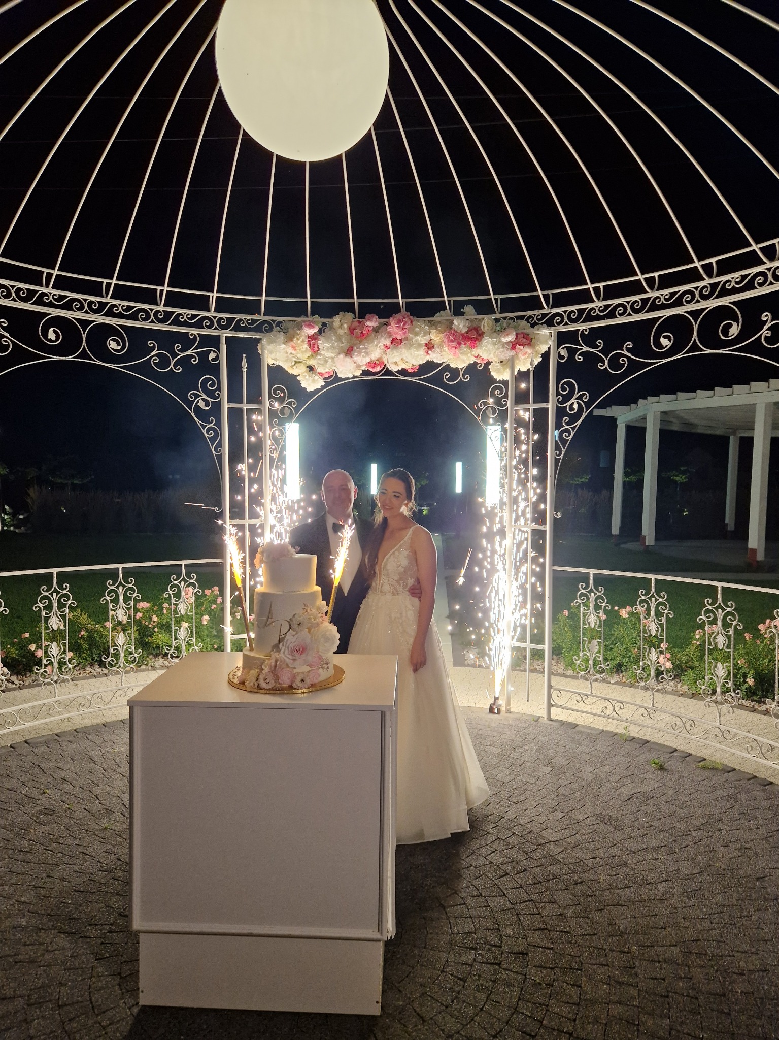 Para młoda pod dekoracyjną altaną z kwiatami i fontannami iskier, obok tort weselny. Uroczysta atmosfera, nocne oświetlenie.