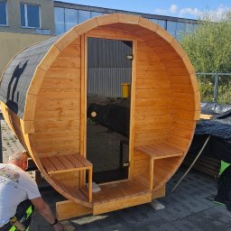 PPHU ROCOSPA - Drewniana sauna w kształcie beczki z przeszklonymi drzwiami, ustawiona na kostce brukowej. Mężczyzna w tle, prawdopodobnie podczas montażu. Dach pokryty gontem.