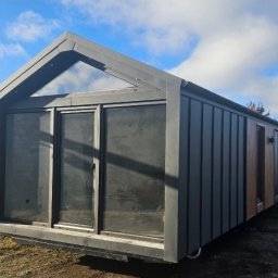 PPHU ROCOSPA - Nowoczesny dom modułowy z ciemnoszarą elewacją i dużymi oknami, widoczny z boku na tle błękitnego nieba z chmurami. Minimalistyczny design z trójkątnym oknem w szczycie.