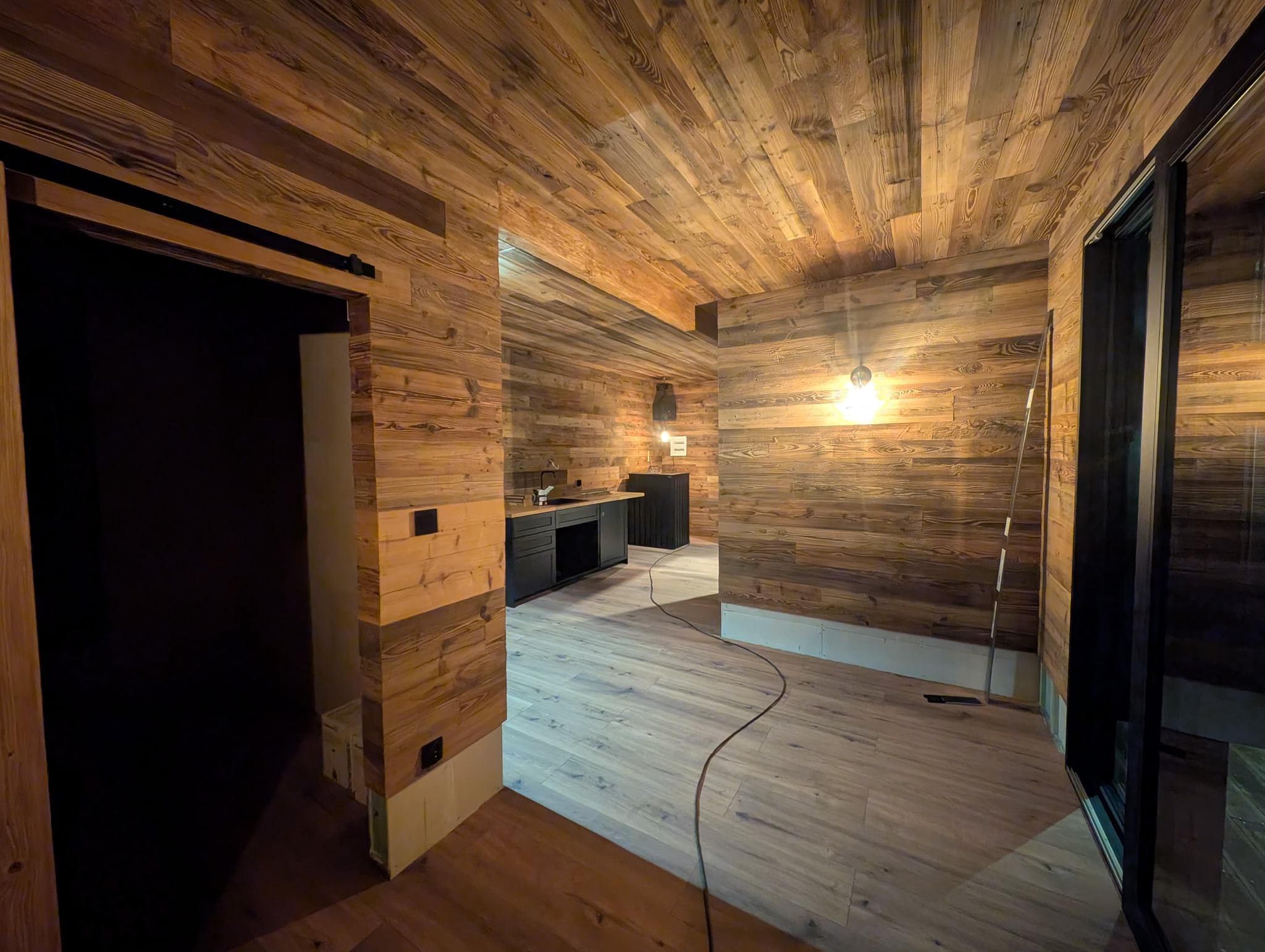 Wnętrze wykończone drewnem: ściany, sufit i podłoga z desek. Widoczna kuchnia z czarnymi szafkami i oknem. Ciepłe oświetlenie podkreśla naturalny rysunek drewna.