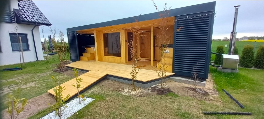 Nowoczesna sauna zewnętrzna z drewnianym tarasem i piecem, otoczona zielenią. Minimalistyczny design z ciemnymi elementami kontrastuje z jasnym drewnem.
