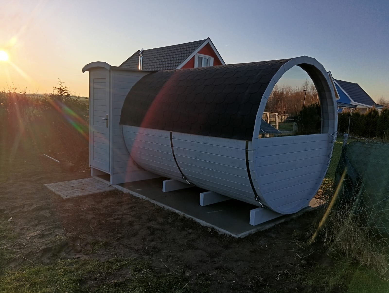 Drewniana sauna w kształcie beczki z przylegającą szatnią, ustawiona na betonowej podstawie na trawniku, w tle domy i zachodzące słońce.