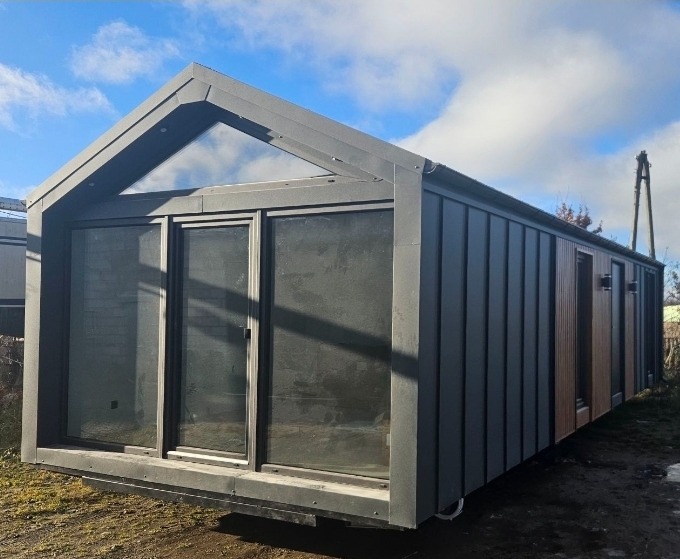 Nowoczesny dom modułowy z ciemnoszarą elewacją i dużymi oknami, widoczny z boku na tle błękitnego nieba z chmurami. Minimalistyczny design z trójkątnym oknem w szczycie.