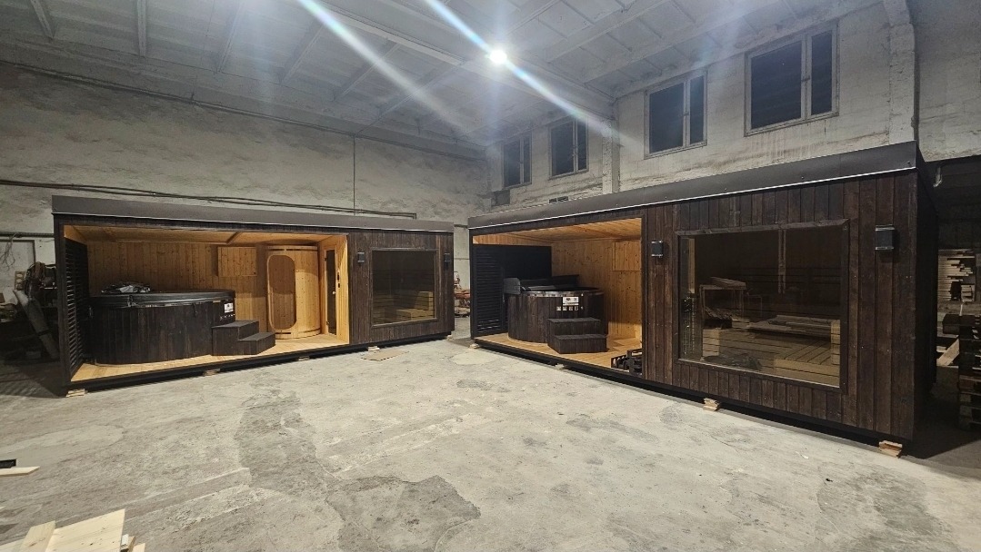 Dwie mobilne sauny z bali z jacuzzi i piecem, ustawione w hali produkcyjnej. Widoczne drewniane wykończenie wnętrza i okna. Oświetlenie górne, surowy beton na podłodze.