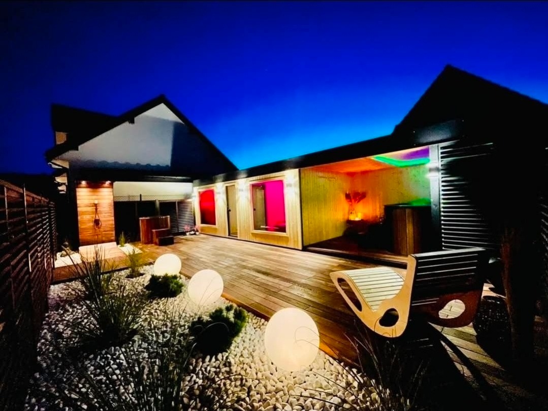 Nowoczesna sauna zewnętrzna z oświetleniem LED i drewnianym tarasem, otoczona białymi kamieniami i dekoracyjnymi kulami świetlnymi, widok nocą.