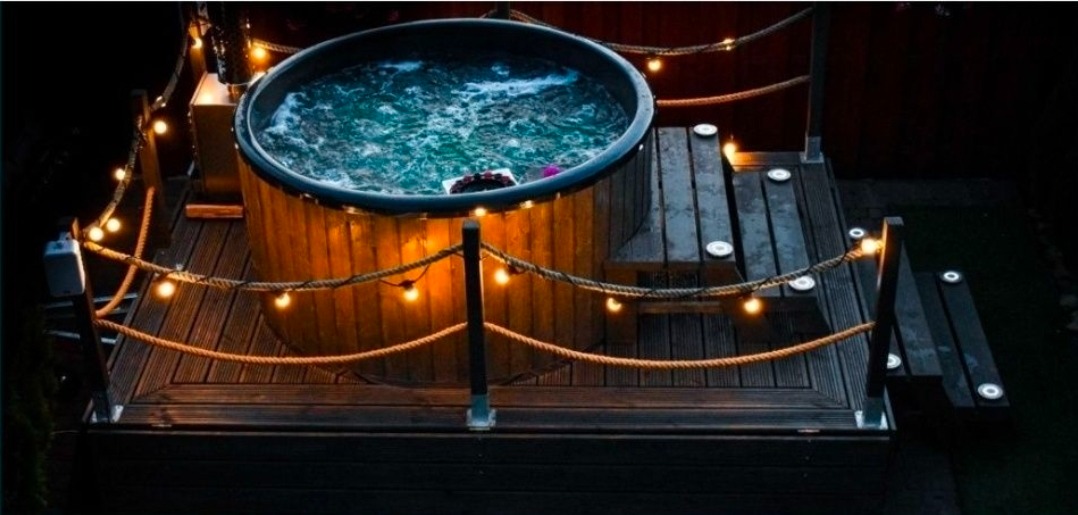 Oświetlona balia ogrodowa z hydromasażem, otoczona drewnianym tarasem i sznurem lampek, tworząca relaksującą atmosferę wieczorową.