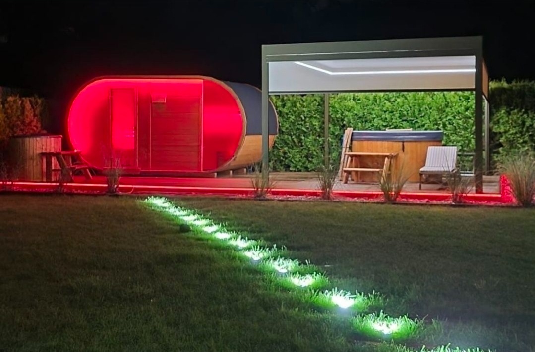 Owalna sauna beczka z czerwonym podświetleniem LED, jacuzzi pod zadaszeniem i ścieżka z lamp ogrodowych na trawniku nocą. Nowoczesny design.