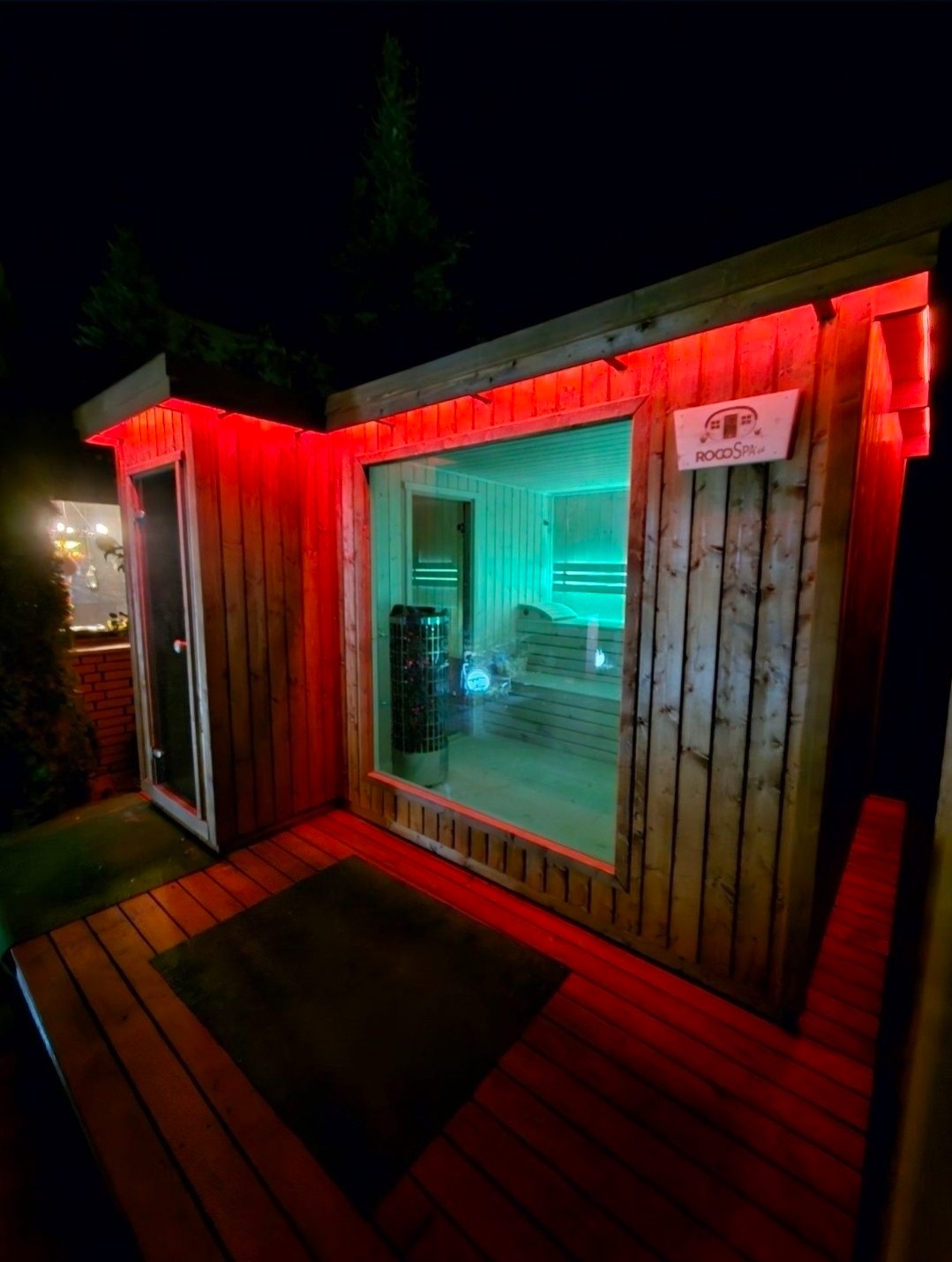 Nowoczesna sauna z bali, oświetlona czerwonymi i zielonymi neonami, widoczna z zewnątrz nocą. Drewniany taras i logo producenta na ścianie.