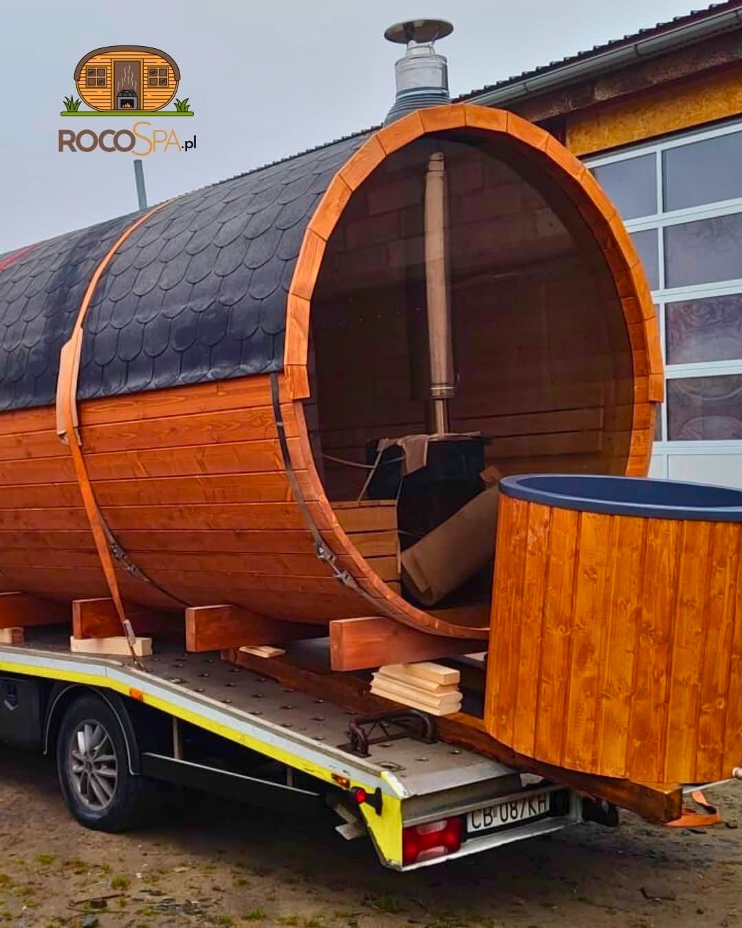 Sauna beczka z piecem i balią na przyczepie transportowej, widok z boku. Dach pokryty gontem bitumicznym. Widoczny komin i logo firmy.