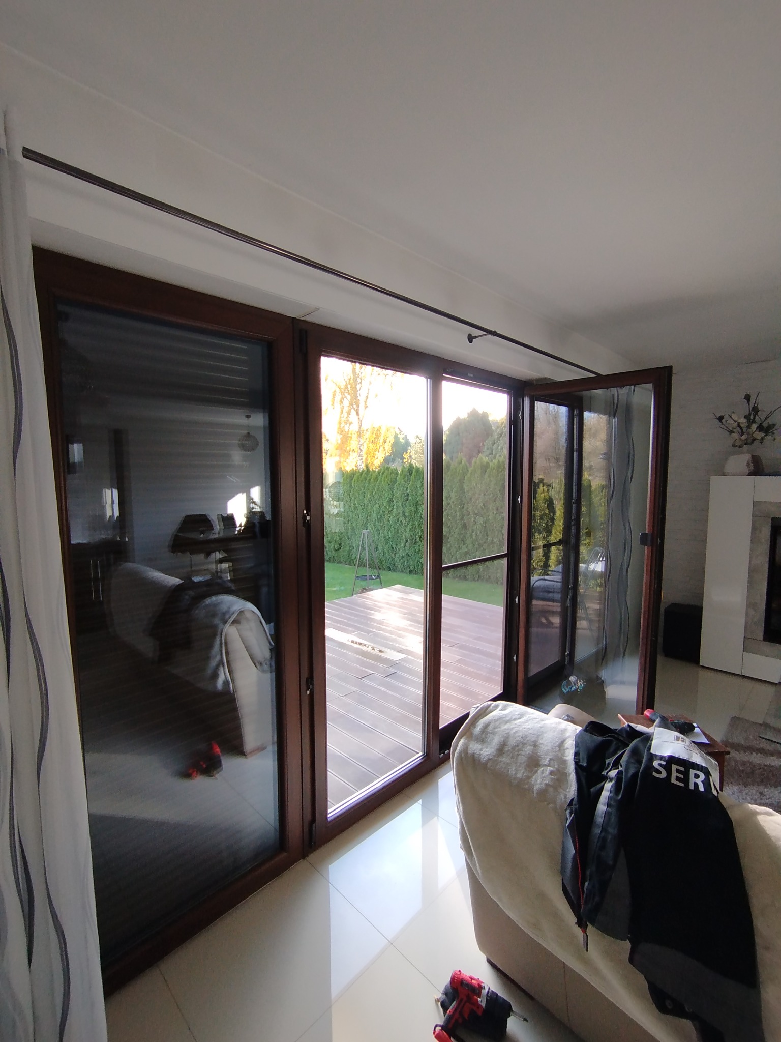 Rozsuwane okno balkonowe z brązowymi ramami, częściowo otwarte na taras z widokiem na ogród. Wnętrze z sofą i zasłoną. Widoczne narzędzia.