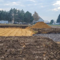 STARBUD Wojciech Starzak - Wyrównany teren pod budowę z wykopem wypełnionym piaskiem, obok hałda piasku i utwardzony fragment drogi. W tle domy i drzewa, z boku koparka i samochód.