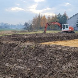 STARBUD Wojciech Starzak - Pomarańczowy koparka Atlas 1304 na placu budowy, przygotowany teren pod budowę, widoczne ślady opon na piasku, w tle budynek i drzewa.