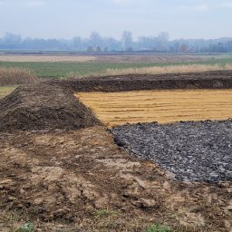 STARBUD Wojciech Starzak - Etap prac ziemnych: wykop z warstwą piasku i kamieni, w tle krajobraz polny i koparka Atlas. Widoczne ślady opon na piasku.