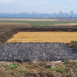 STARBUD Wojciech Starzak - Etap prac ziemnych: warstwa żwiru, piasku i obrzeże z ziemi na tle pola uprawnego i zamglonego horyzontu. Widoczny fragment koparki.