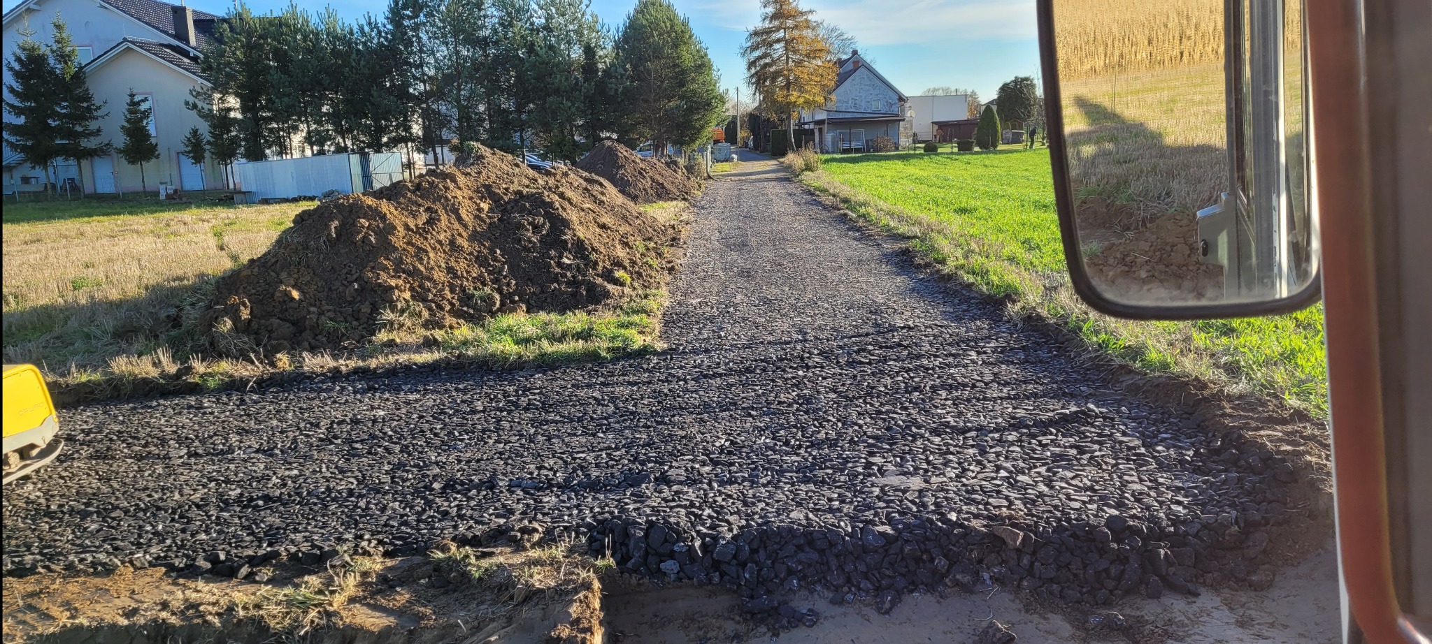 Utwardzona droga z nasypami ziemi po bokach, widok z kabiny maszyny. Kamienie tworzą równą powierzchnię. W tle domy i drzewa.