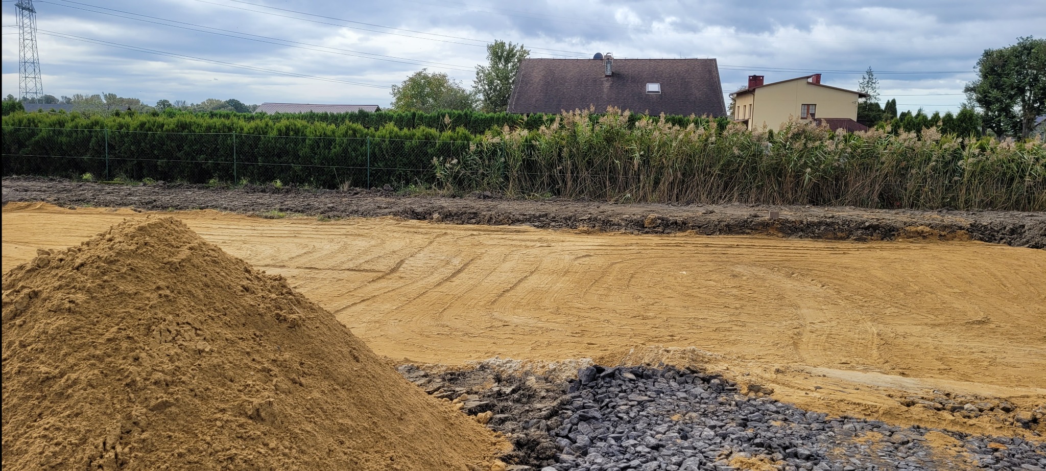 Przygotowany teren pod budowę z nasypanym piaskiem i kamieniami, widoczne ślady po maszynach, w tle zabudowania i zieleń.