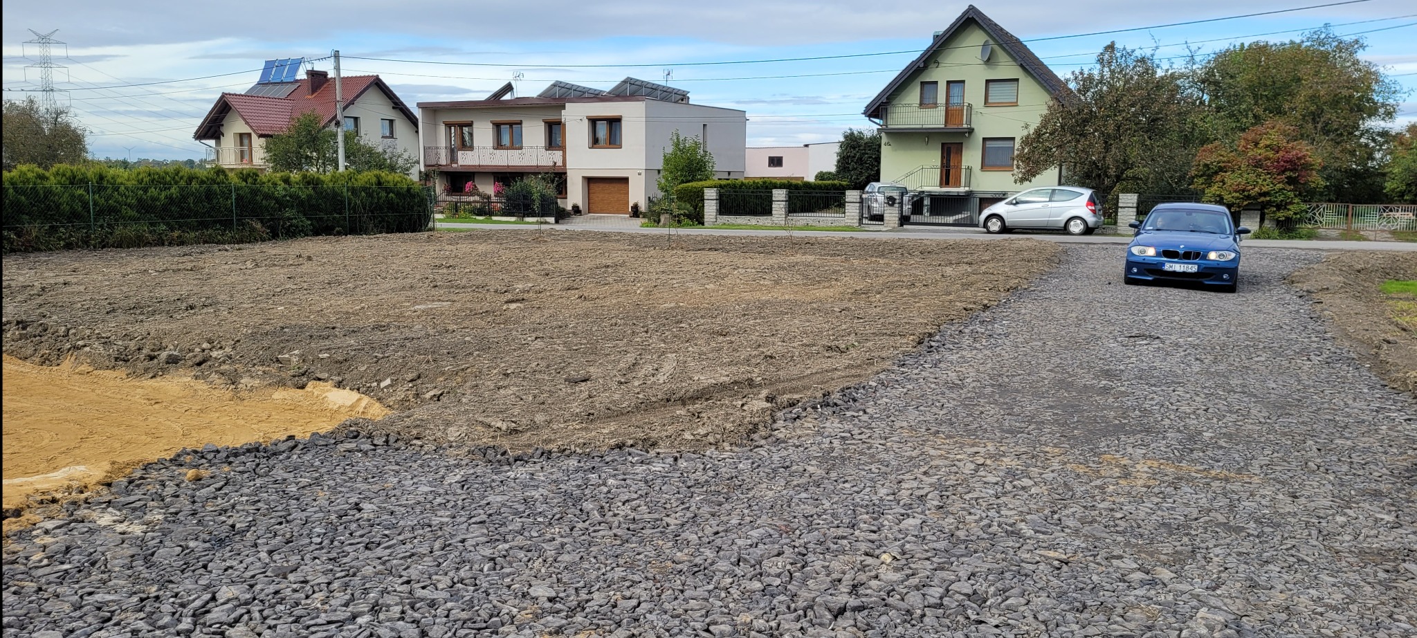 Przygotowany teren pod budowę domu z utwardzoną drogą dojazdową z kamienia. W tle zabudowa jednorodzinna i błękitne auto.