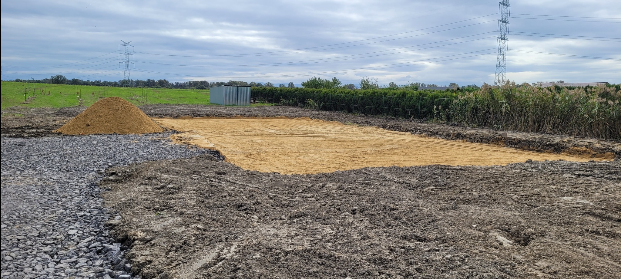 Przygotowany teren pod budowę w Odra: wyrównany piasek, obok hałda piasku i kamieni, w tle zielona łąka i słupy wysokiego napięcia.