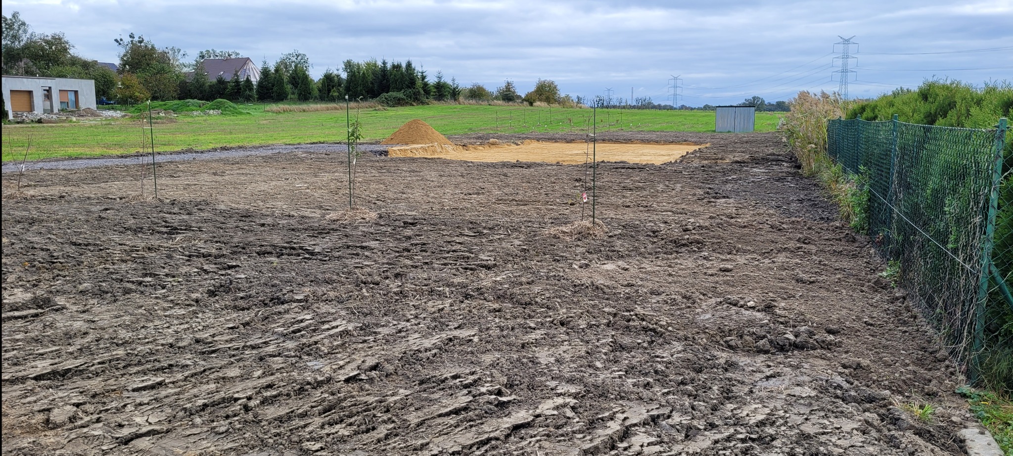 Przygotowany teren pod budowę z wykopem, usypaną górką piasku i młodymi drzewkami. Widoczne ogrodzenie z siatki i słupy wysokiego napięcia w tle.
