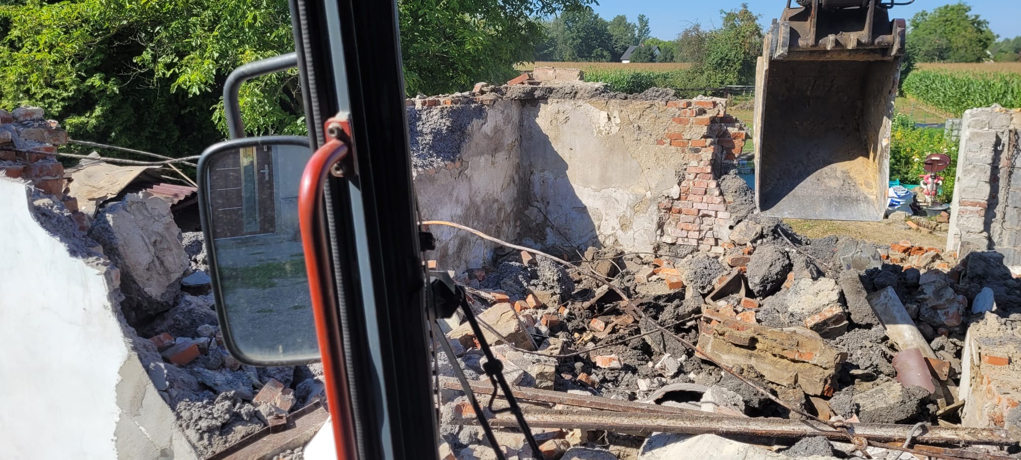 Demontaż ceglanego budynku z perspektywy kabiny koparki. Widoczne gruz, fragmenty ścian i łyżka koparki. W tle zieleń drzew i zabudowania.