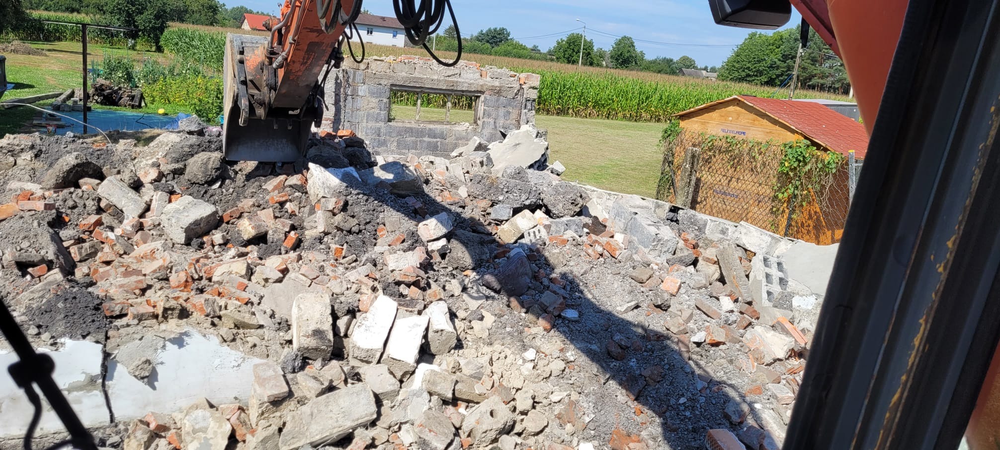 Demolka budynku z użyciem koparki. Gruz ceglany i betonowy na pierwszym planie, w tle pole kukurydzy i budynek gospodarczy. Widok z kabiny operatora.