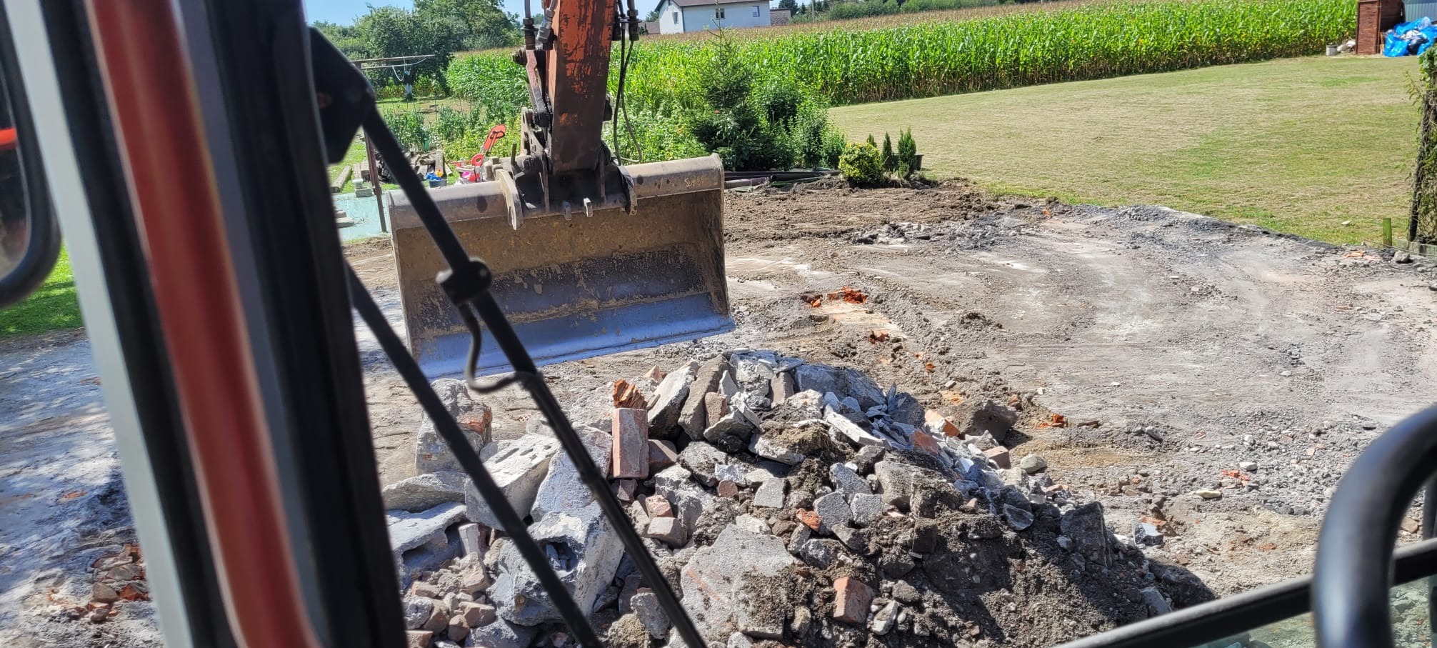 Pomarańczowa koparka z łyżką nad stertą gruzu na placu budowy, widok z kabiny. W tle zielone pole i dom.