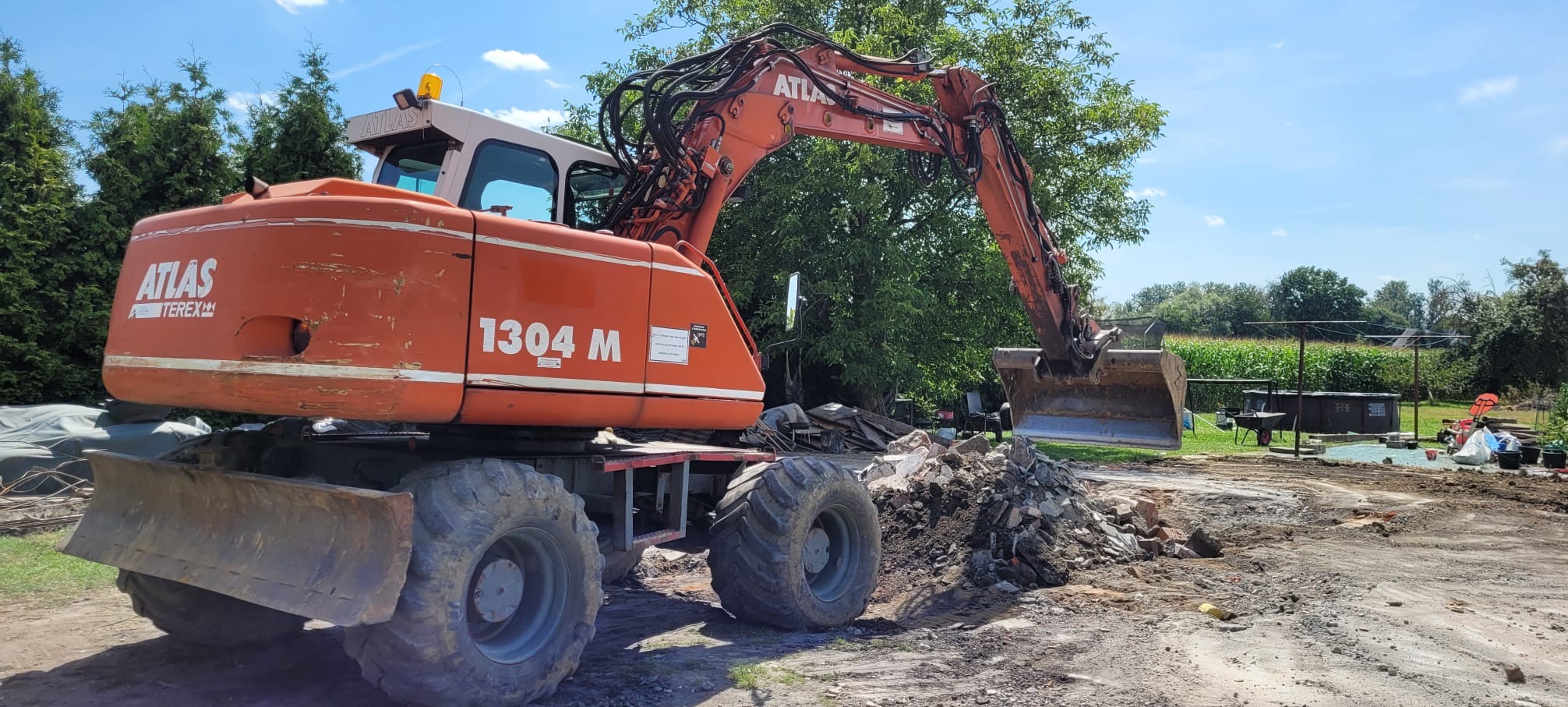 Pomarańczowa koparka Atlas 1304 M na kołach, z łyżką nad stertą gruzu na placu budowy, w tle zieleń i błękitne niebo. Prace ziemne w toku.