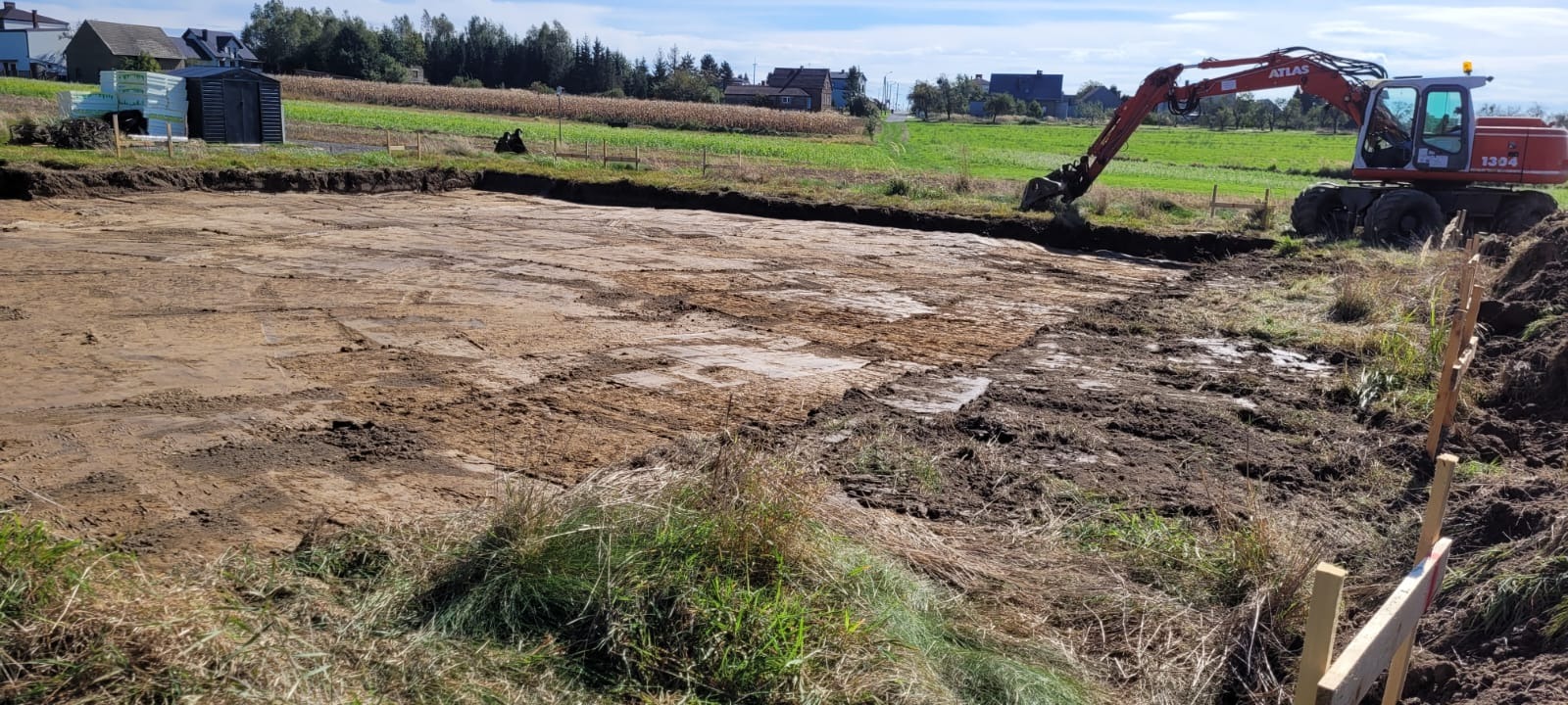 Wyrównany teren pod budowę z widocznym wykopem i pracującą koparką Atlas 1304 w tle, otoczony zielenią i zabudowaniami.