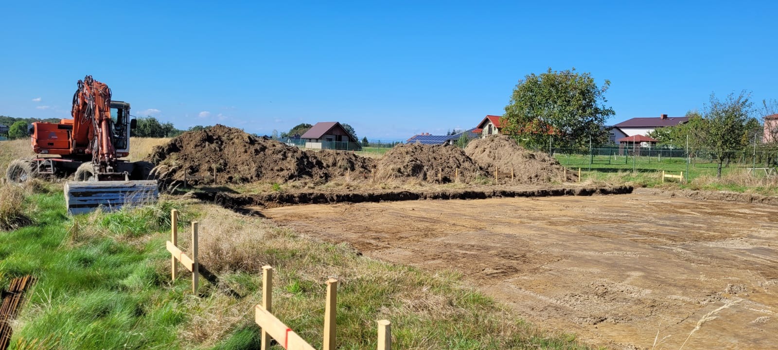 Wykop pod fundamenty domu jednorodzinnego, z hałdami ziemi i pomarańczową koparką w tle, w słoneczny dzień. Widoczne drewniane paliki wyznaczające obrys.