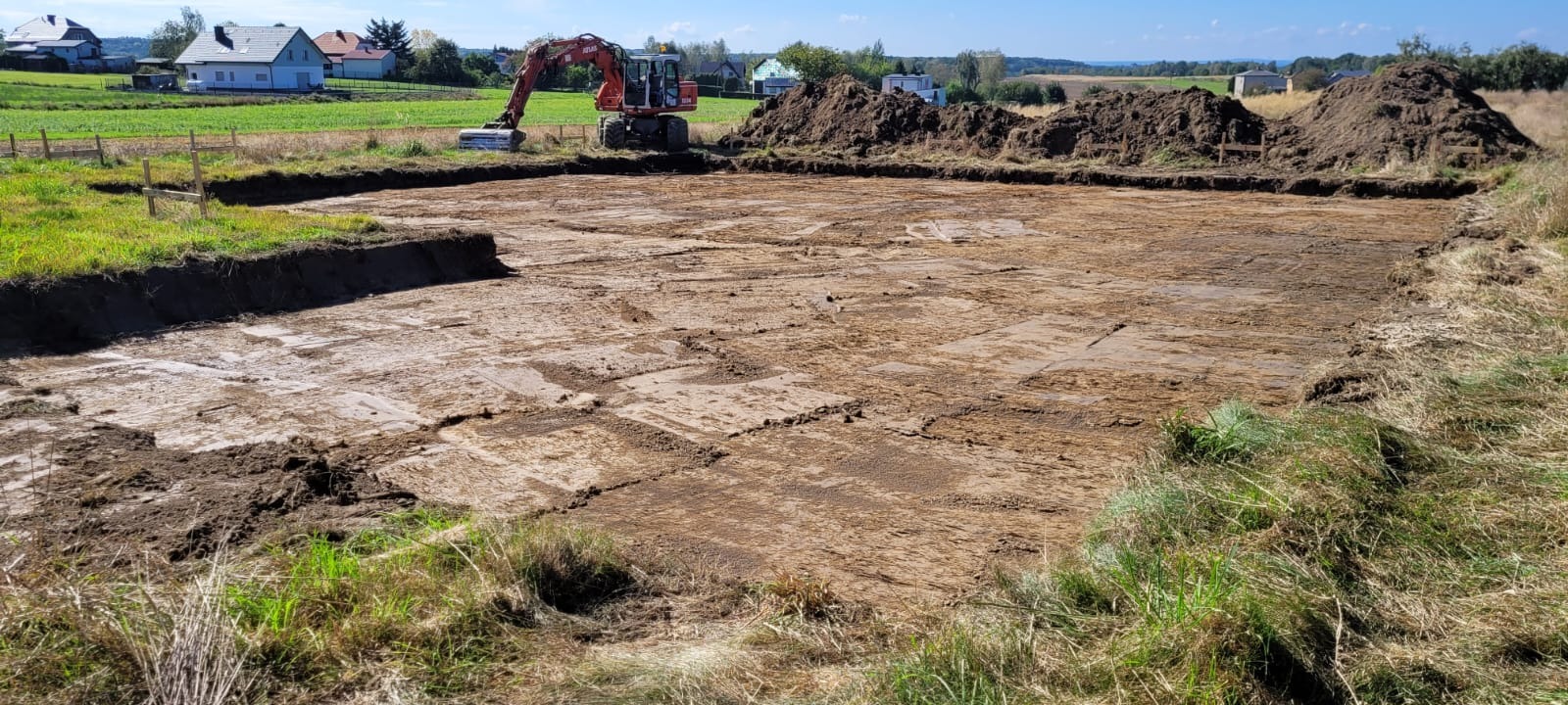Wykop pod fundamenty z widocznym wykopanym terenem, stertą ziemi i koparką w tle, w słoneczny dzień. Widoczne drewniane znaczniki.