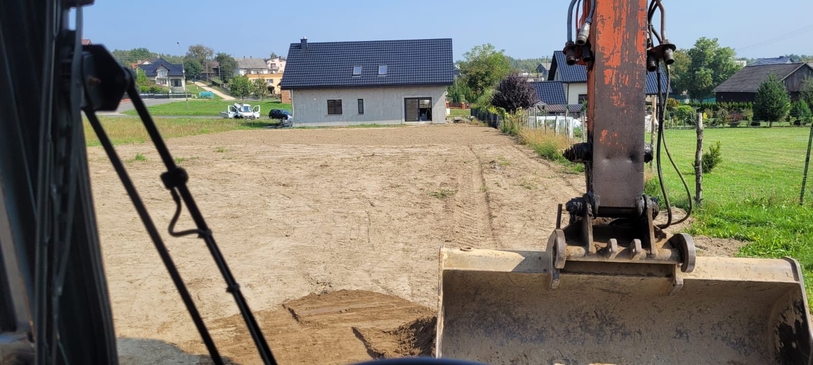 Widok z kabiny koparki na przygotowany teren pod budowę domu jednorodzinnego. W tle dom w stanie surowym zamkniętym i inne zabudowania.