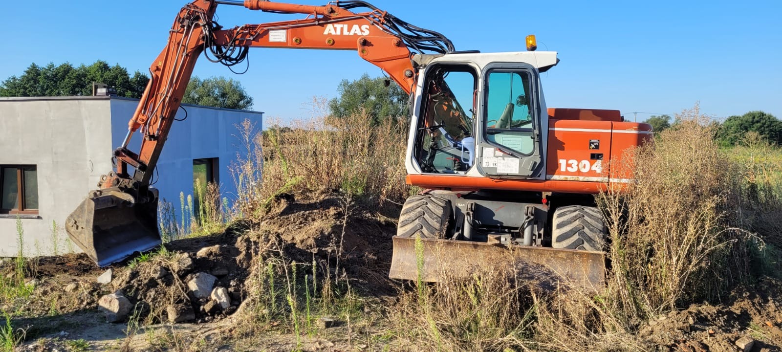 Pomarańczowa koparka Atlas 1304 w trakcie prac ziemnych przy budynku. Widoczny wykop i pryzma ziemi. Tło stanowi błękitne niebo i zieleń drzew.