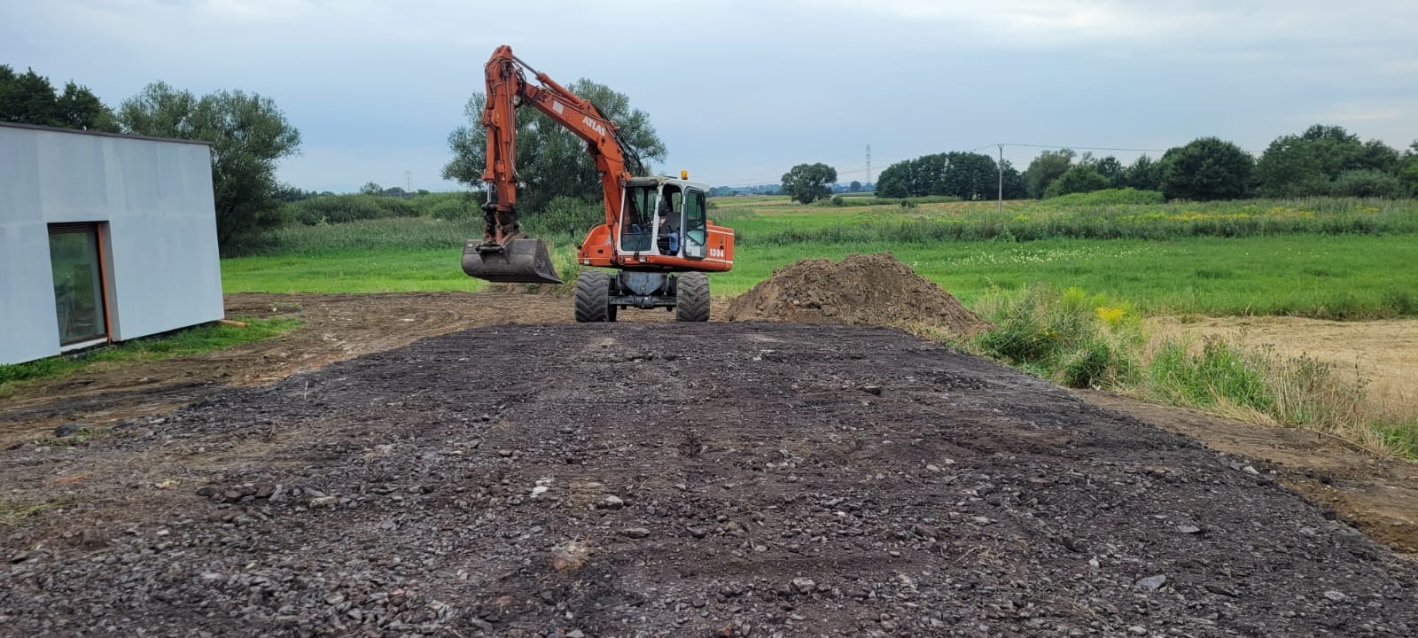 Pomarańczowa koparka Atlas na tle zielonego pola i nowego budynku. Wykopy ziemne, niwelacja terenu. Prace ziemne w toku.