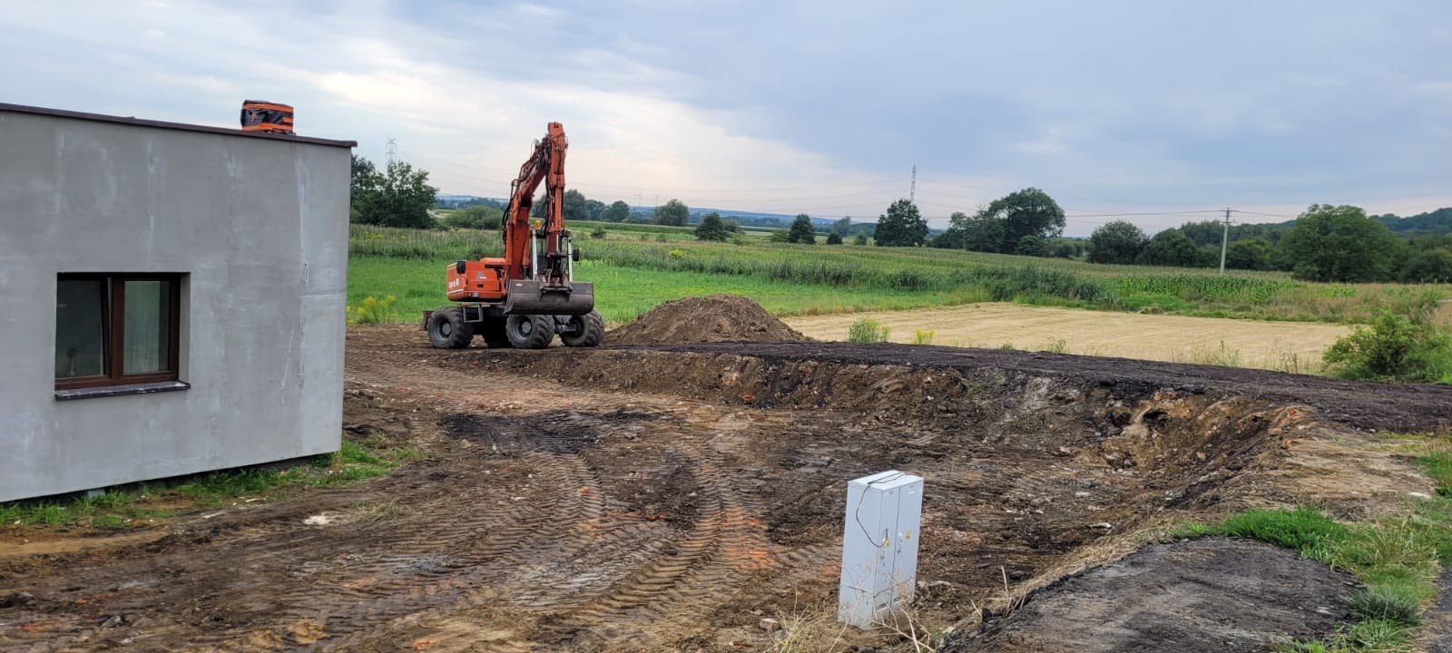 Pomarańczowa koparka kołowa na placu budowy obok szarego budynku z brązowymi oknami. W tle zielone pola i drzewa pod pochmurnym niebem.