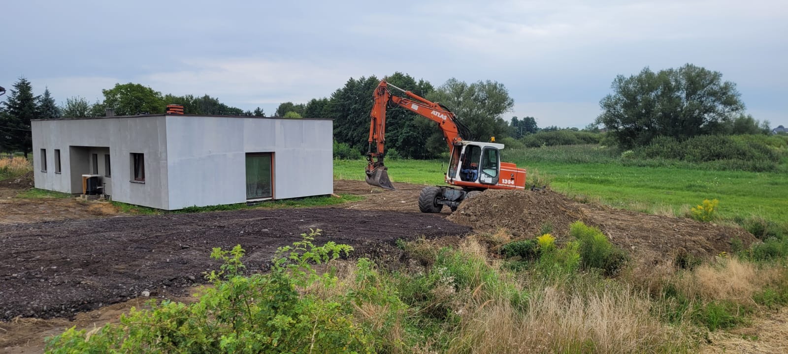 Pomarańczowa koparka Atlas 1304 przy białym, parterowym domu. Przygotowany teren pod budowę, widoczne ślady pracy maszyny. Trawa i drzewa w tle.