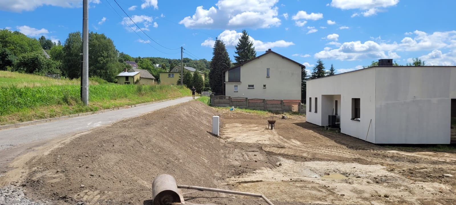 Ukształtowany teren przy drodze i nowym budynku, widoczny walec do ubijania ziemi. Prace ziemne w toku, błękitne niebo z chmurami w tle.