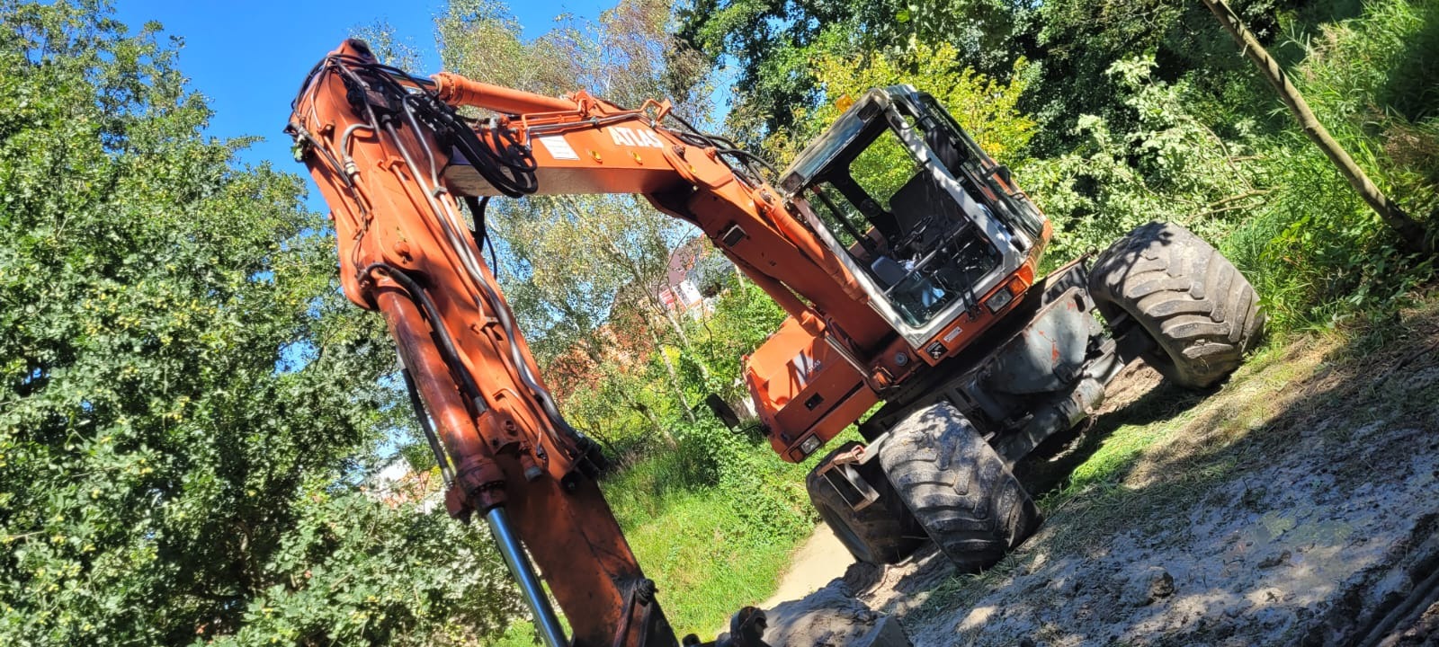 Pomarańczowa koparka kołowa Atlas na błotnistym terenie, otoczona zielenią drzew. Widok z dołu, podkreślający masywność maszyny i jej gotowość do pracy.